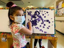 A little girl wearing a facemask while painting on a canvas setup on an easel.