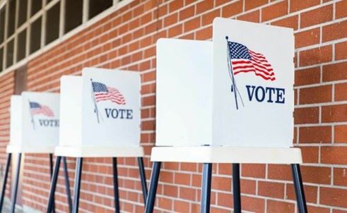 Three white stools lined up against a brick wall. There are folders with the word vote and an American Flag on top of the stools.