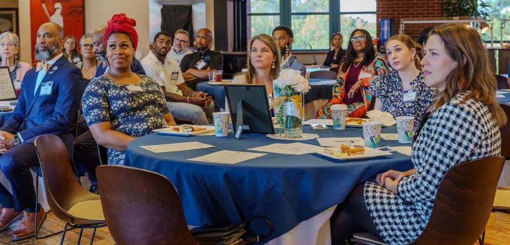 A room full of individuals sitting at dining tables while listening to a speaker.