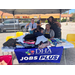 Four individuals sitting behind the DHA Jobs Plus booth that is pilled high with clothes and books.