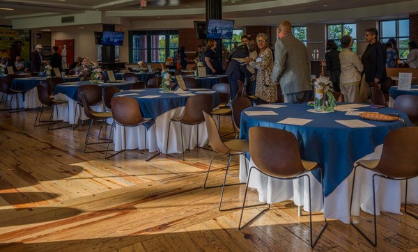 A big room full of individuals mingling around dinning tables.