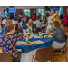 Individuals eating at a dinning table while others greet each other in the background.